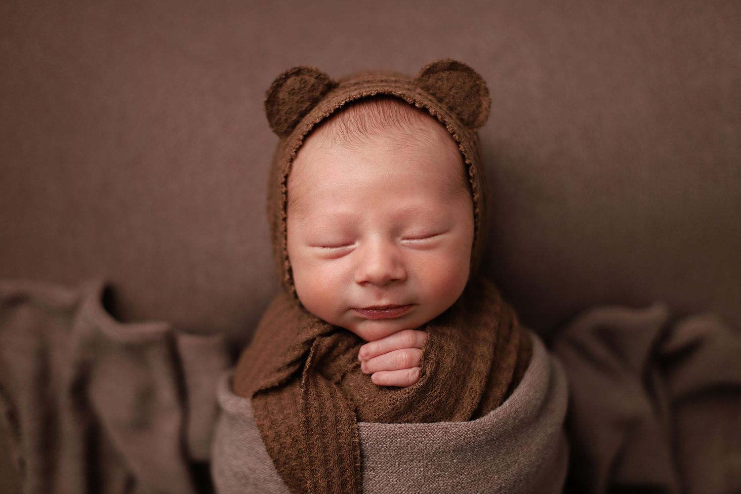 A sleeping baby wearing a brown bear-style bonnet and swaddled in cozy blankets against a dark backdrop. Utah newborn photography