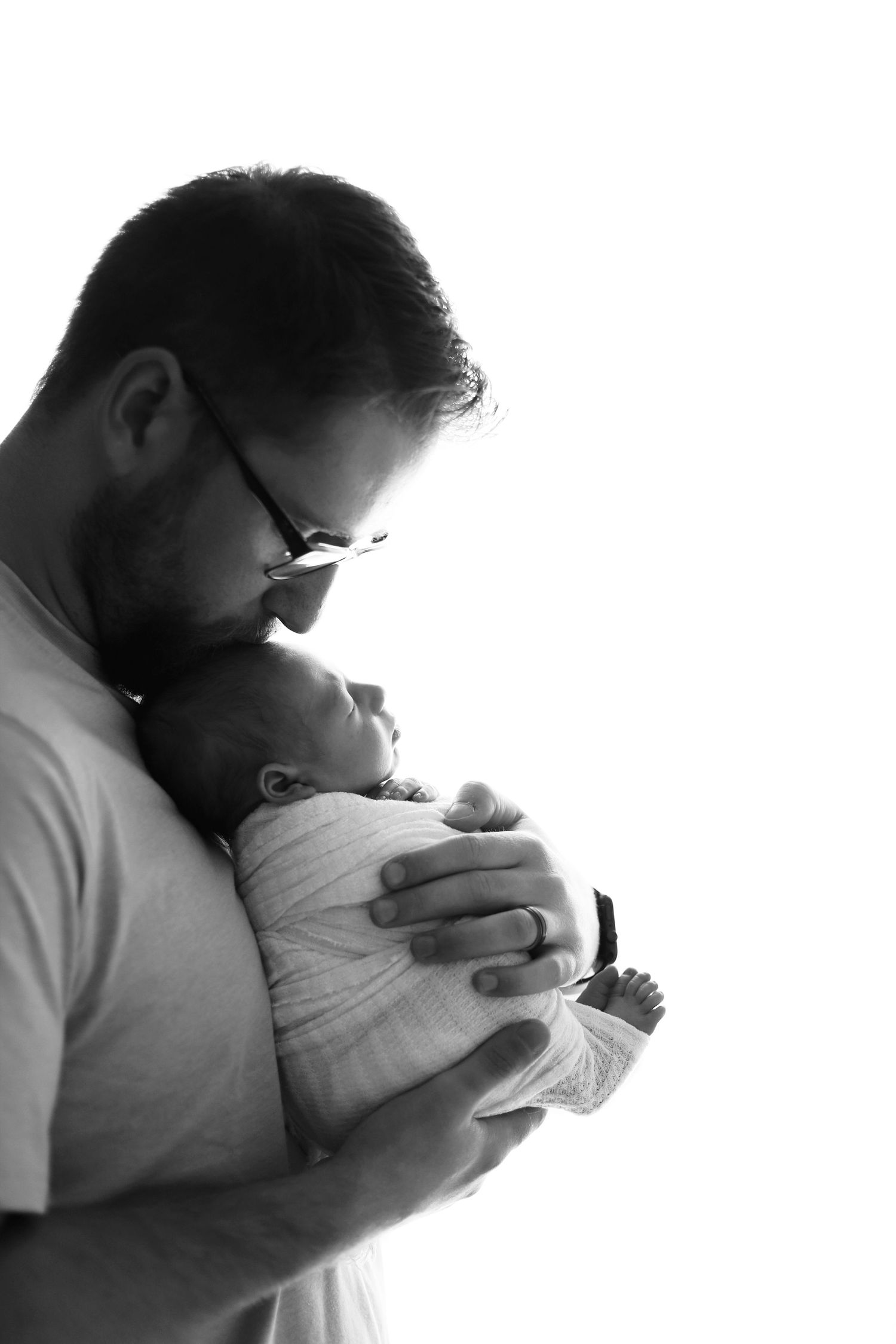 A tender black and white silhouette of a person wearing glasses holding a swaddled newborn baby against their chest. newborn photography