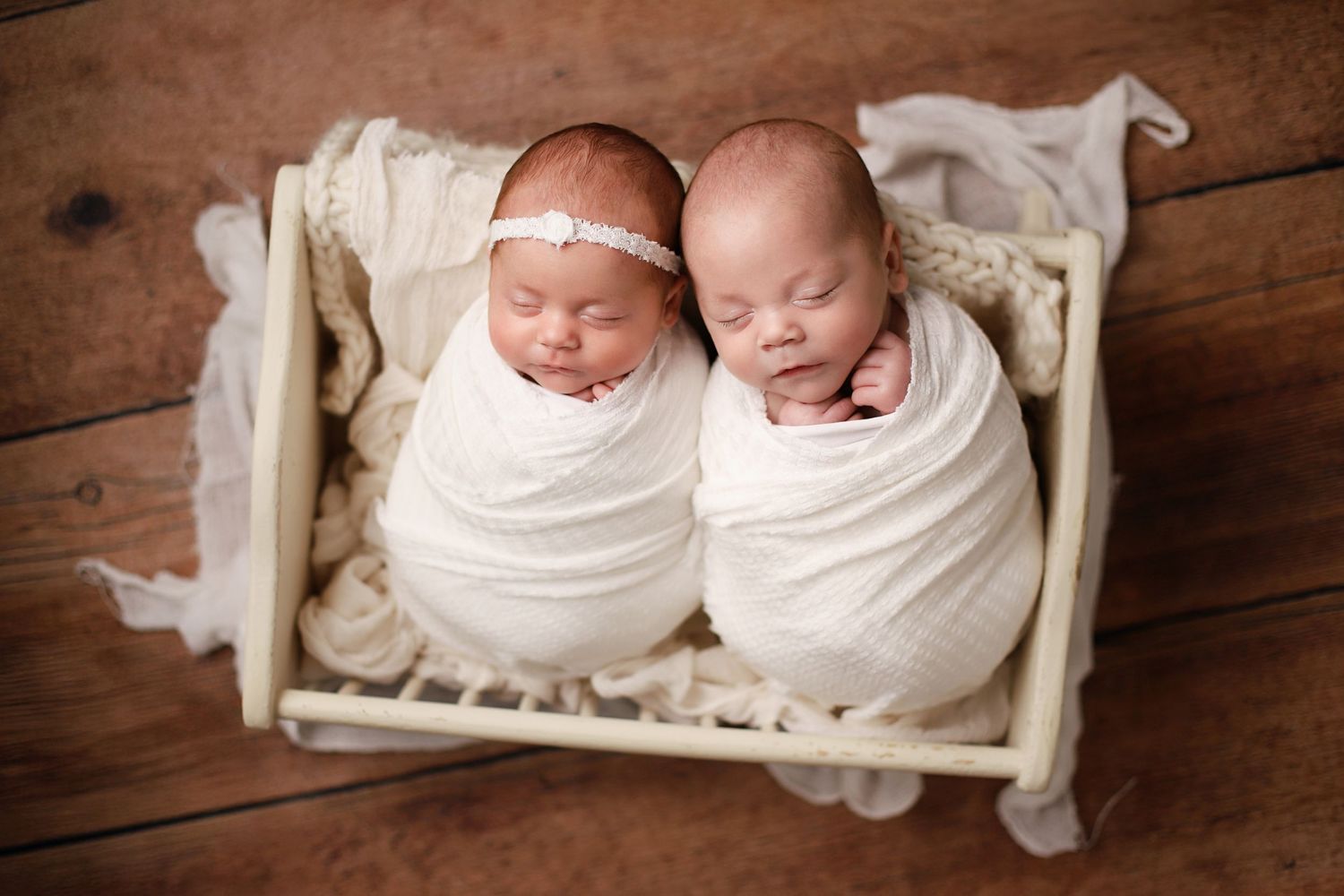 Two swaddled newborns sleeping side by side on a wooden floor and white bedding in matching white wraps. Utah twin newborn photographer