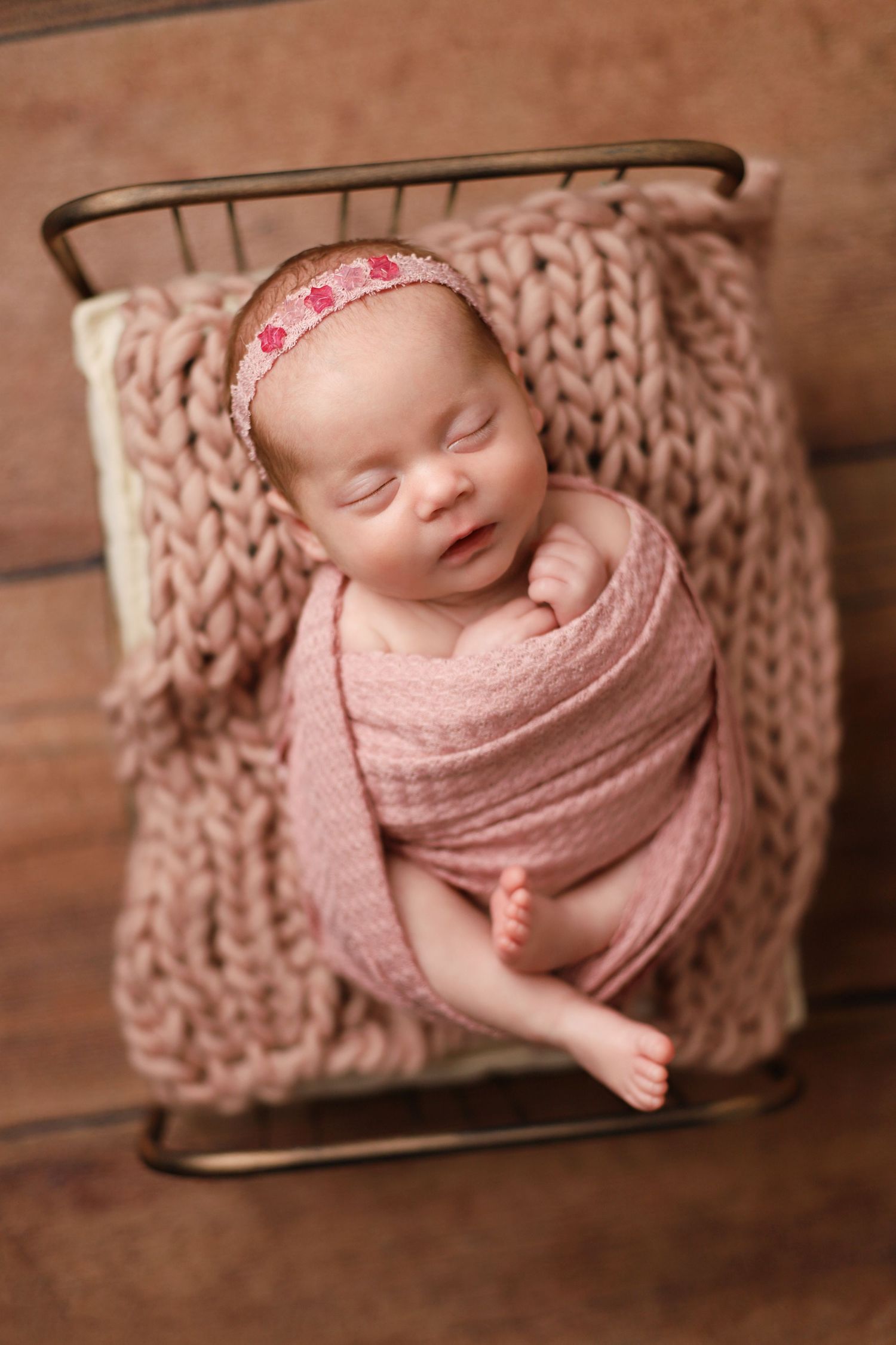 A sleeping newborn wrapped in pink swaddle rests on chunky knit blanket in vintage metal baby bed. Orem newborn photographer