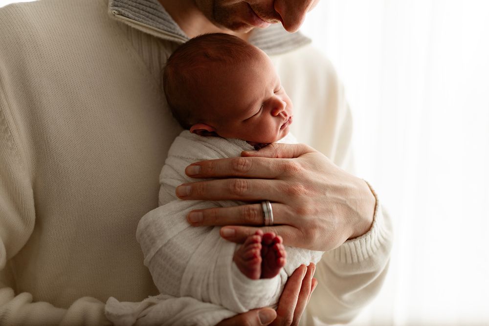 Parent cradling a sleeping newborn baby wrapped in a white swaddle blanket, showing tiny red feet.