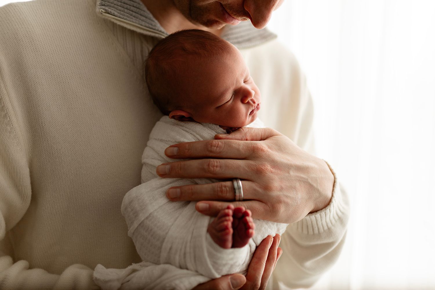 Parent cradling a sleeping newborn baby wrapped in a white swaddle blanket, showing tiny red feet.