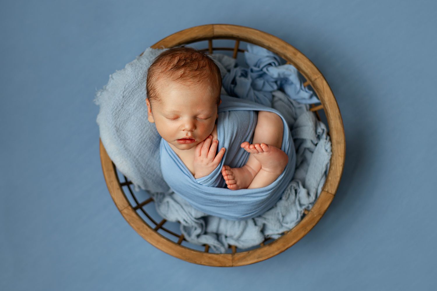 Newborn baby sleeping peacefully in a wooden bowl wrapped in a soft blue cloth on a blue background.