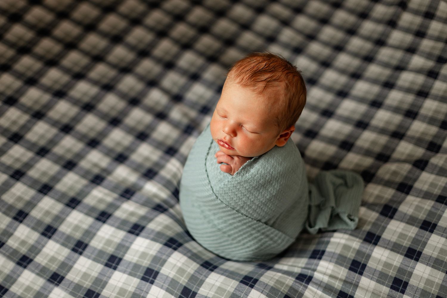 Newborn baby swaddled in a soft blue wrap, sleeping peacefully on a navy plaid blanket.