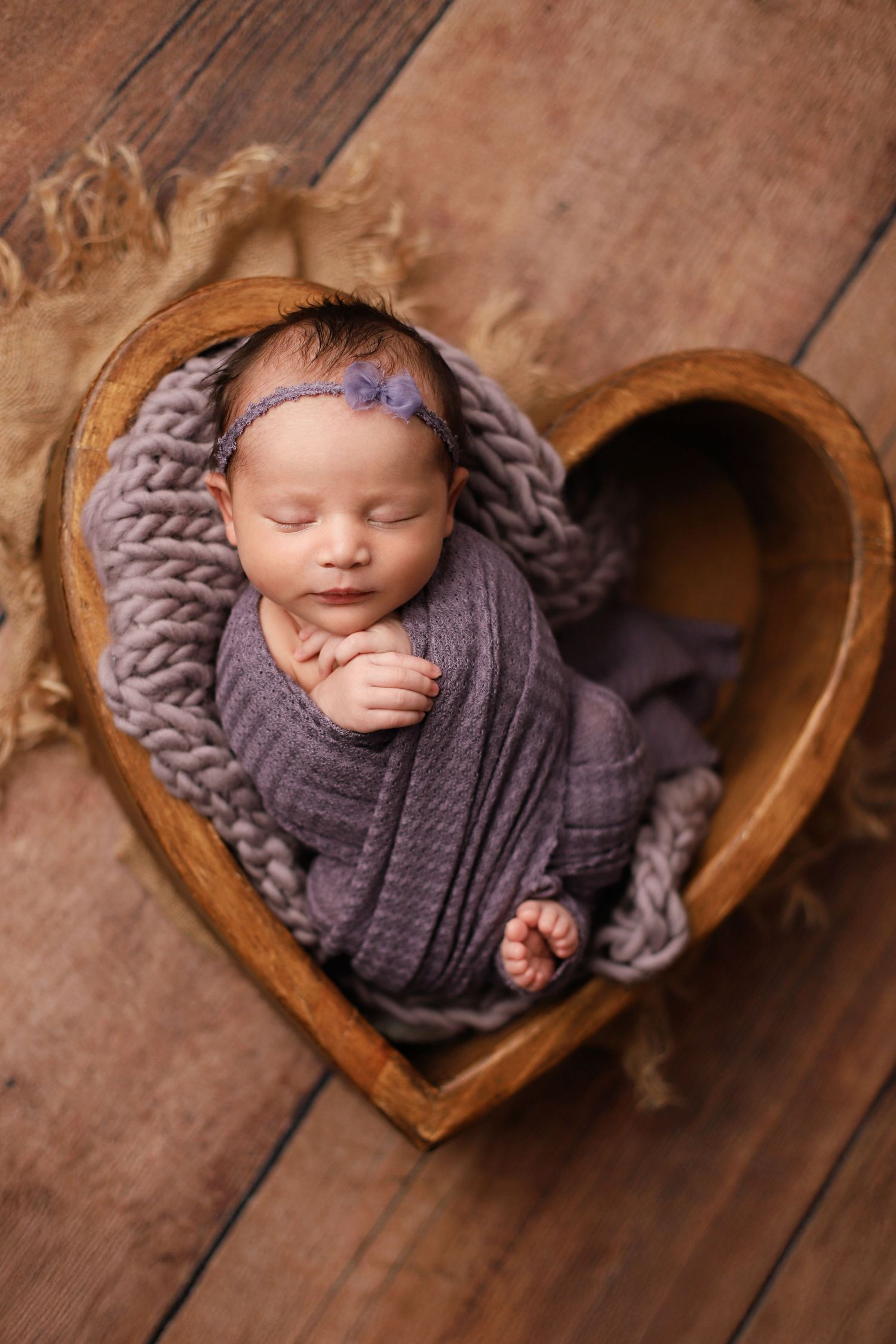 Newborn wrapped in gray knit blanket sleeping peacefully in wooden heart-shaped bowl on rustic floor. Utah County newborn photography