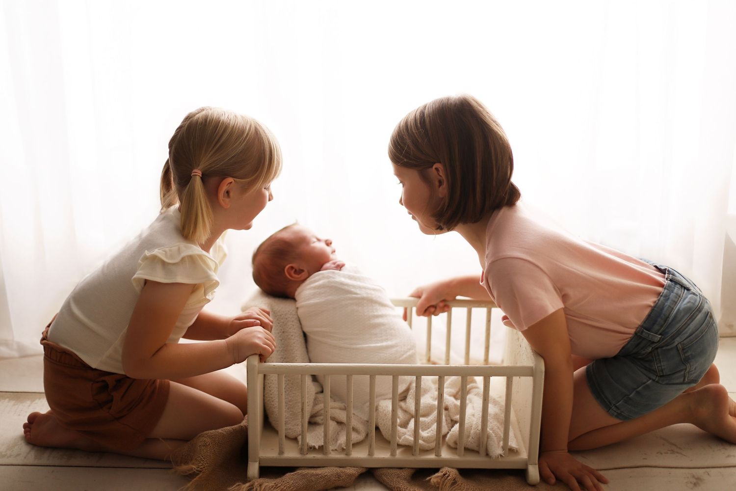 Two young children lean over to look at a baby lying in a white crib next to a bright window. newborn and sibling photography