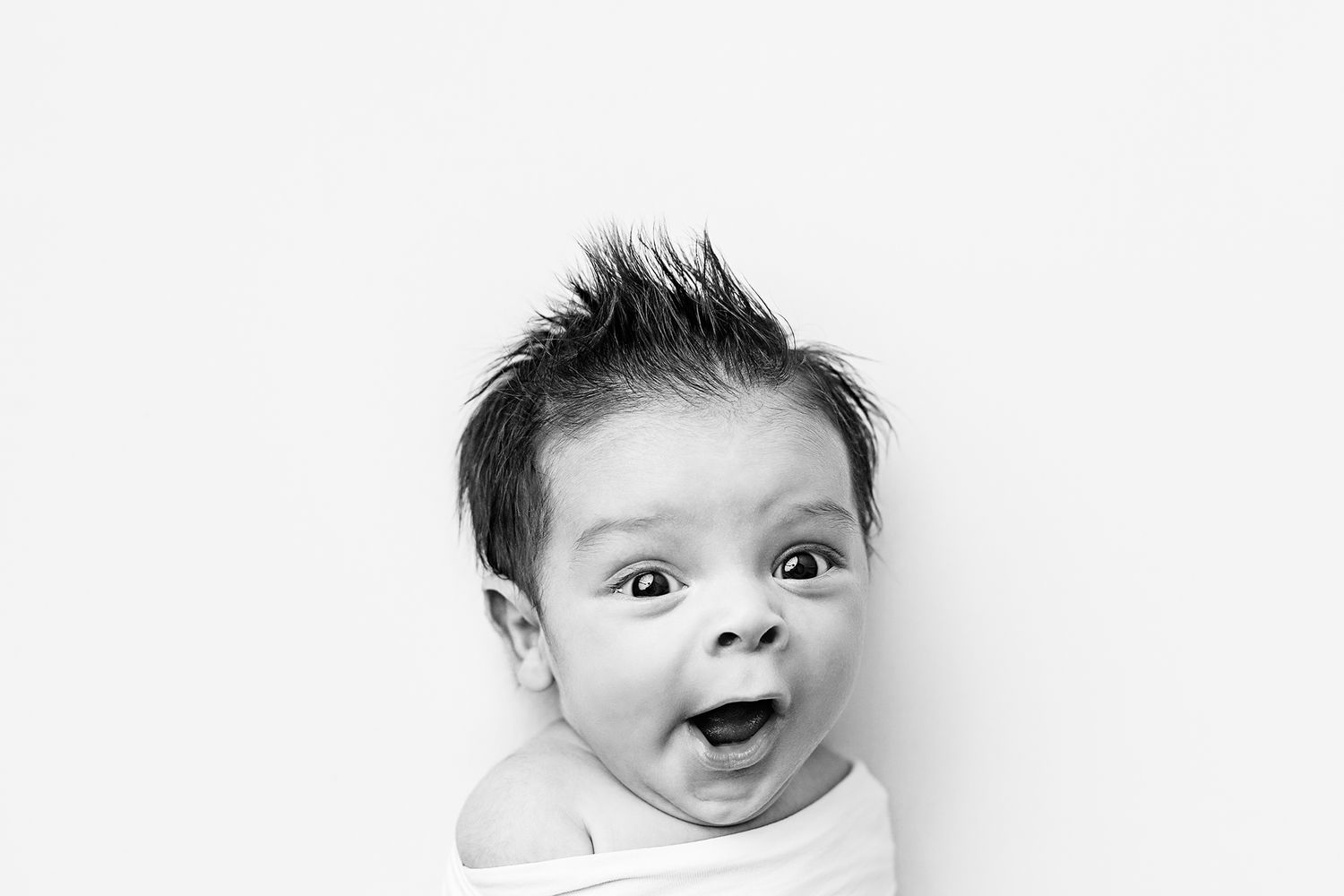 Black and white photo of a surprised newborn baby with spiky hair, wide eyes, and open mouth, wrapped in white.