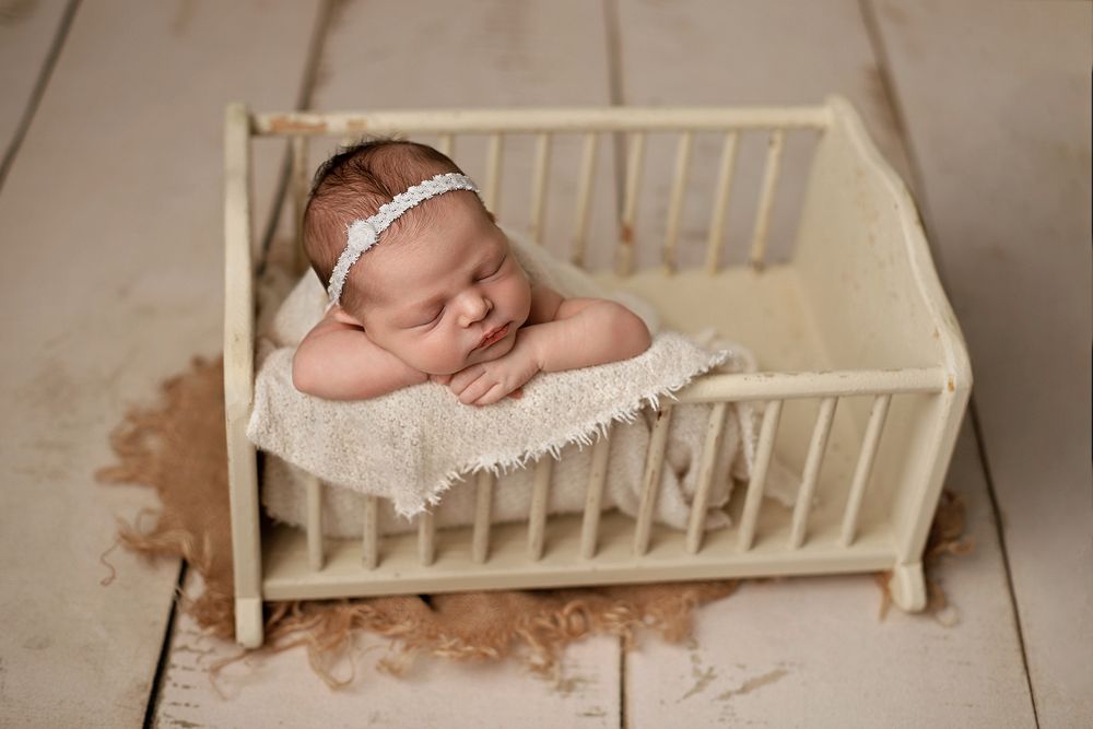 Newborn baby sleeping peacefully in a tiny wooden crib with lace headband during professional photo session.