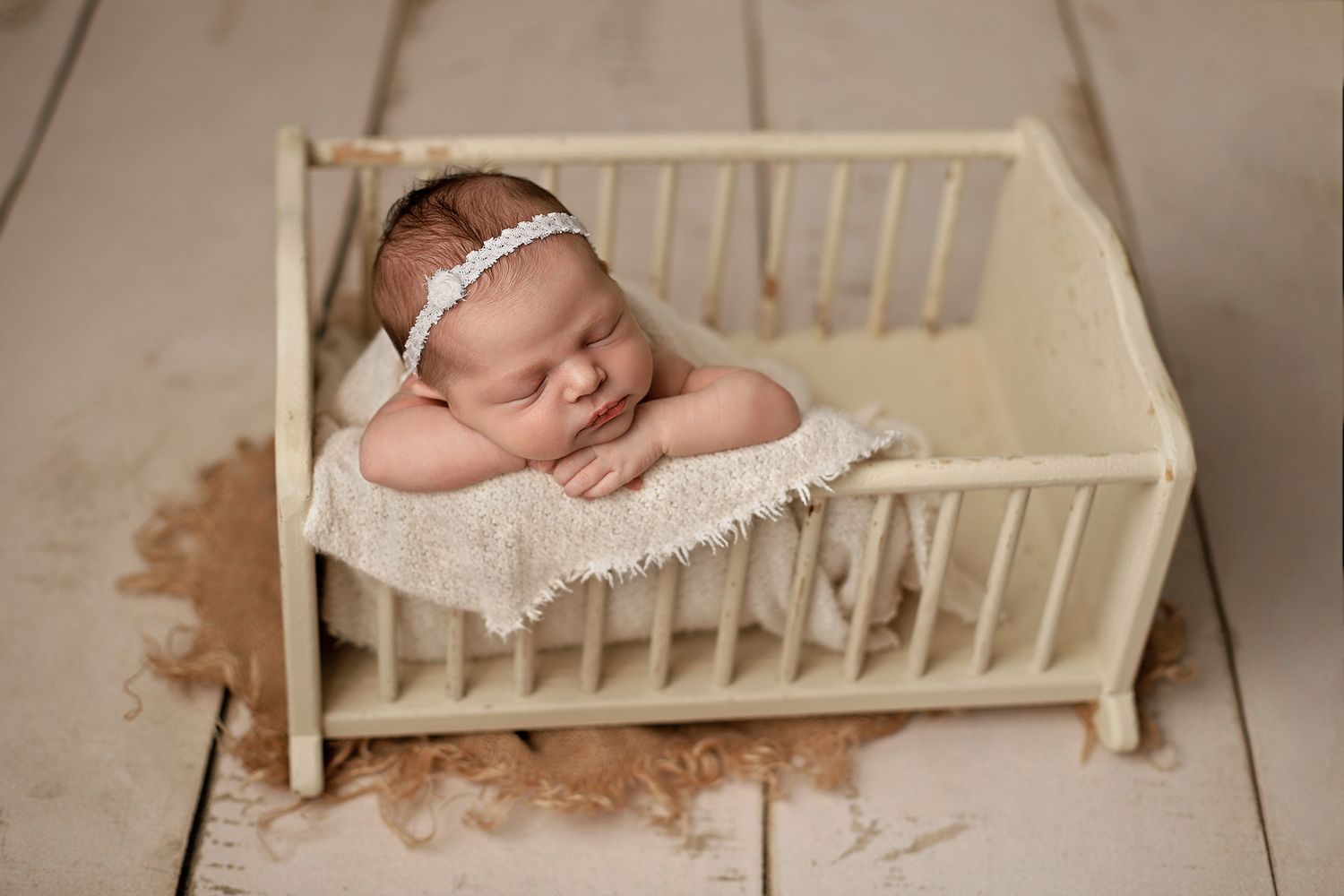 Newborn baby sleeping peacefully in a tiny wooden crib with lace headband during professional photo session.