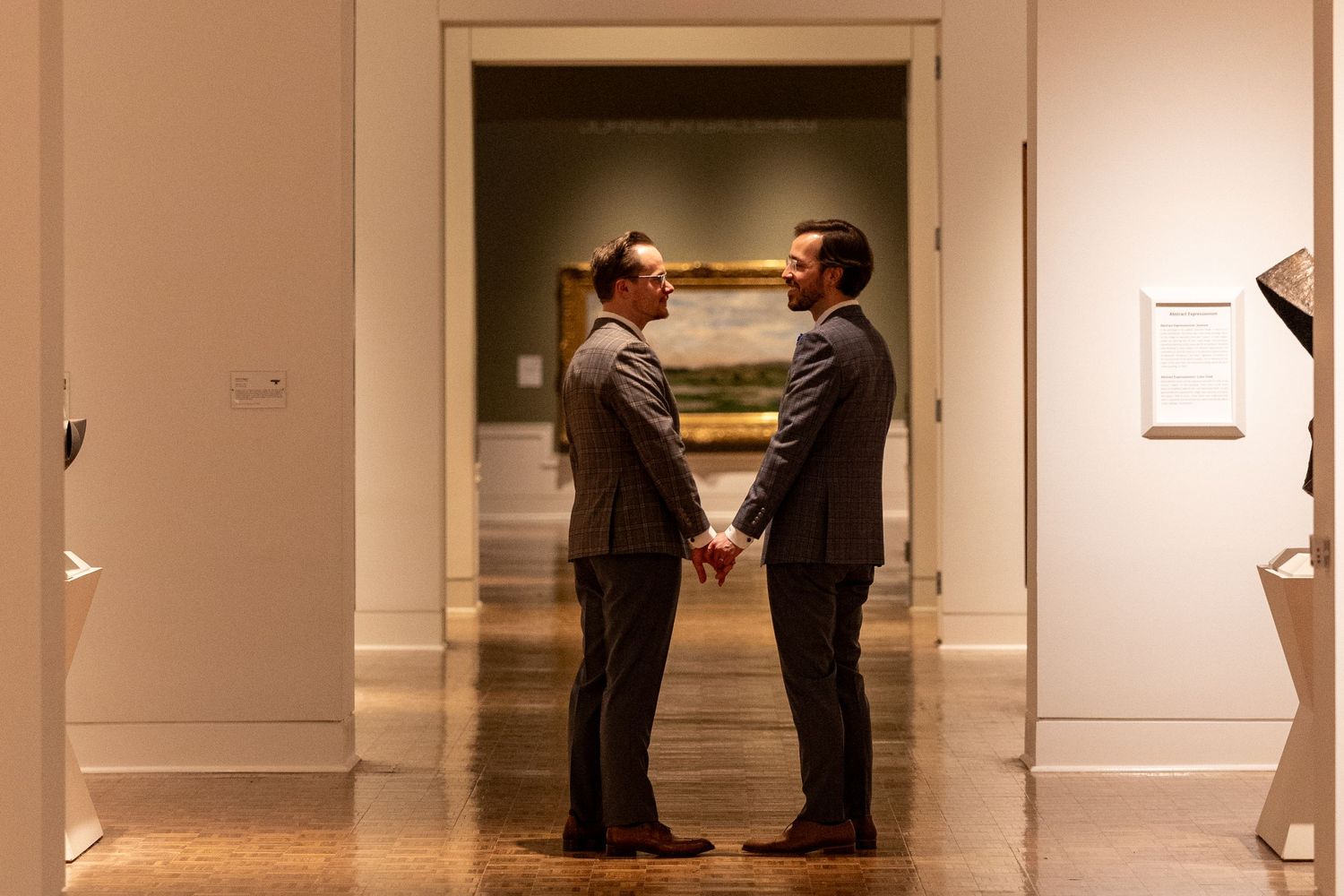 Two grooms in suits hold hands facing each other in an art museum gallery during their wedding.