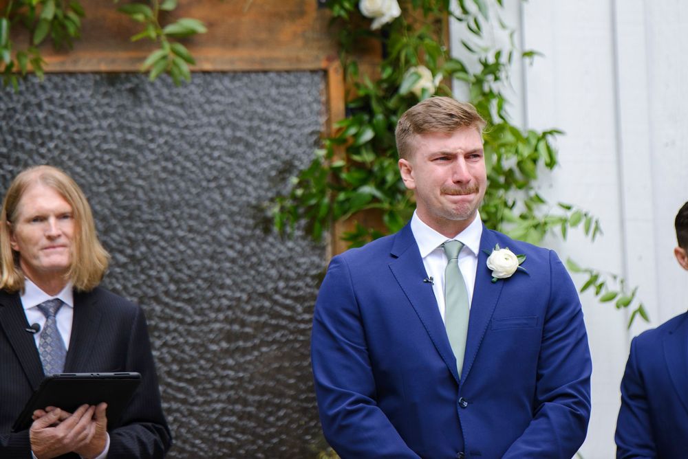 A groom in a navy blue suit with a mint green tie stands next to a wooden wall and leafy green vines.