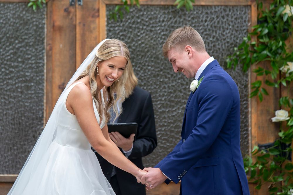 A bride and groom exchange rings during an outdoor wedding ceremony surrounded by stone walls and greenery.