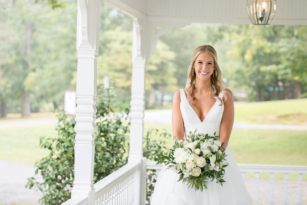 A person in a white dress stands on an elegant white porch with decorative green floral arrangements.