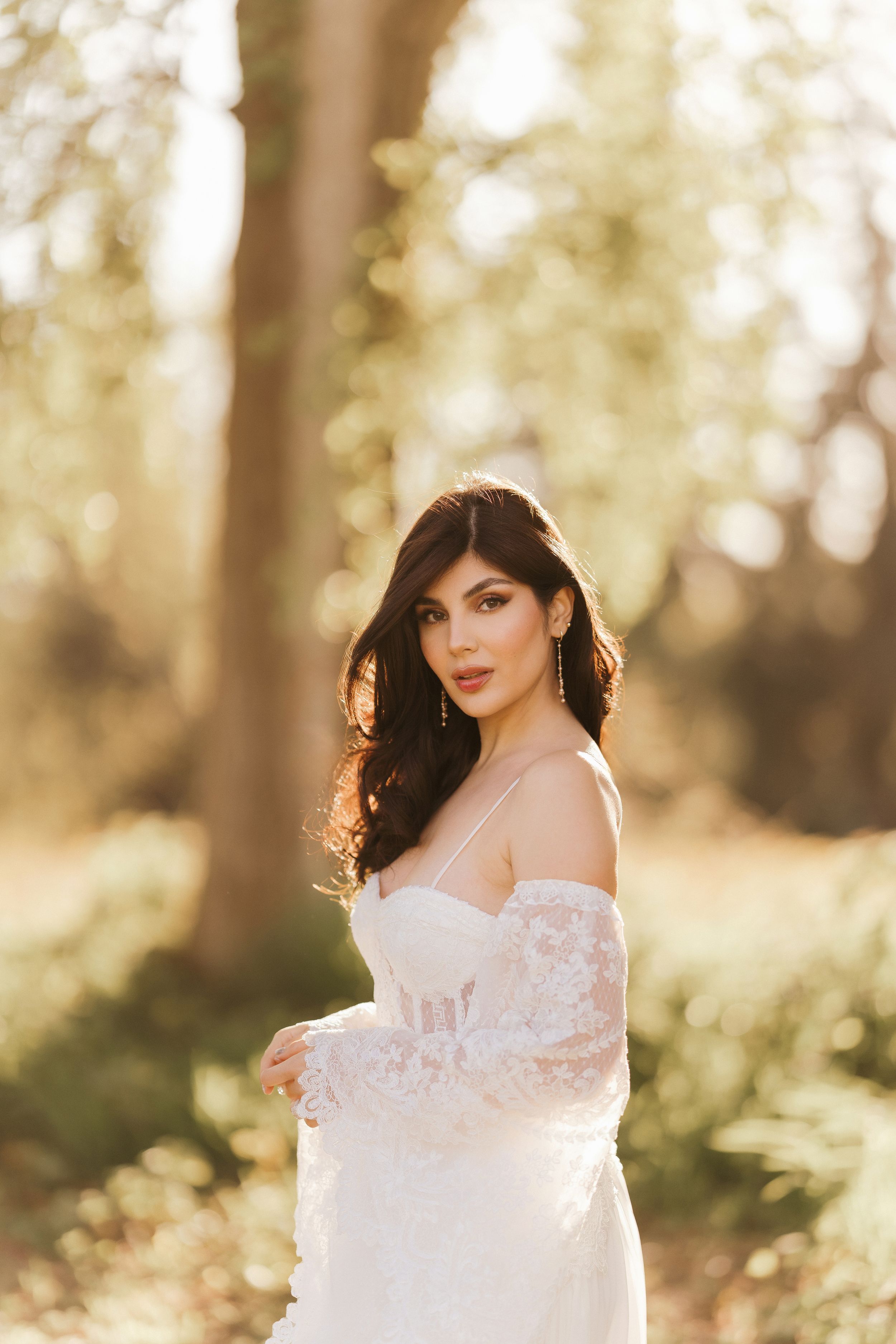 A woman in a white lace gown poses gracefully in a sun-drenched forest setting during a bridal photoshoot.