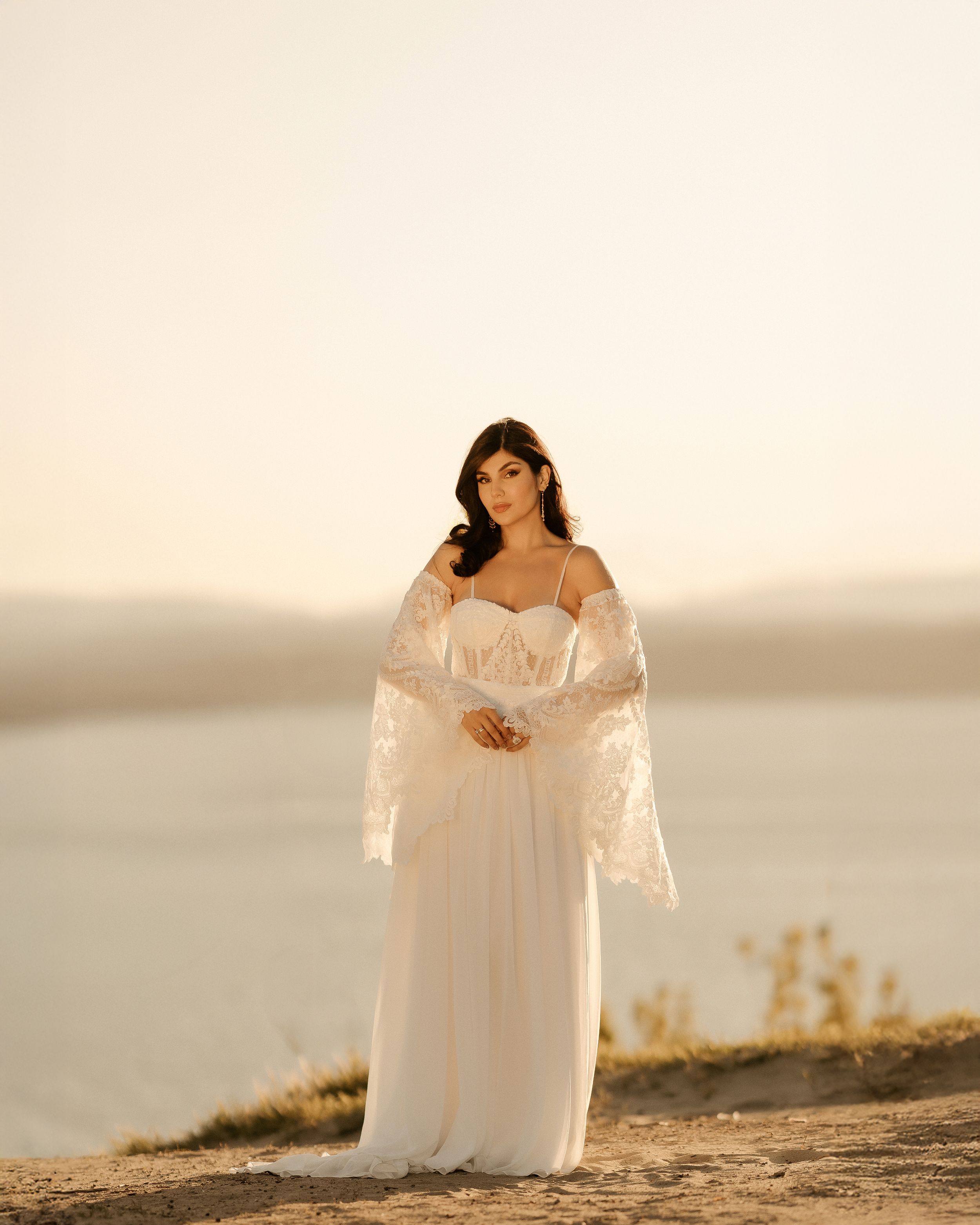 A person in a flowing white wedding dress stands gracefully against a serene sunset backdrop on a sandy hill.