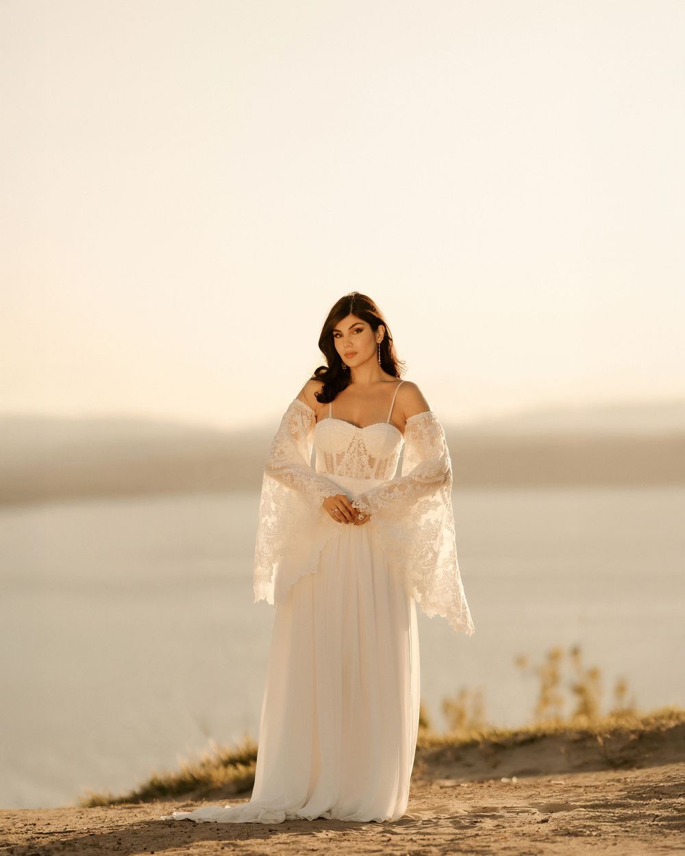 A person in a flowing white wedding dress stands gracefully against a serene sunset backdrop on a sandy hill.