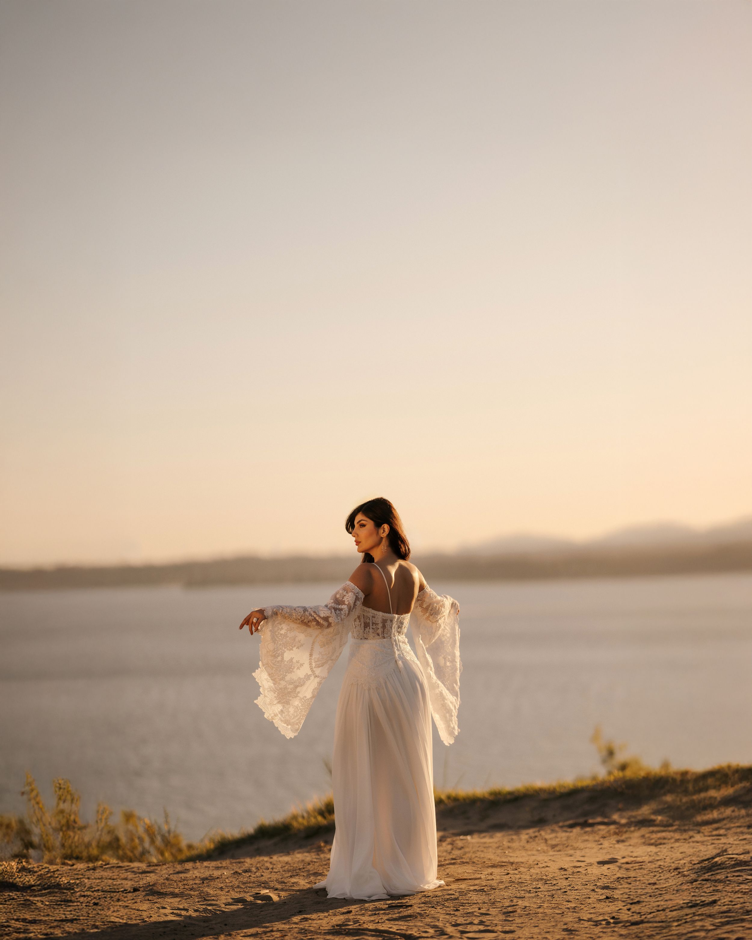 A person in a flowing white dress stands gracefully on a cliff overlooking a serene lake at sunset.