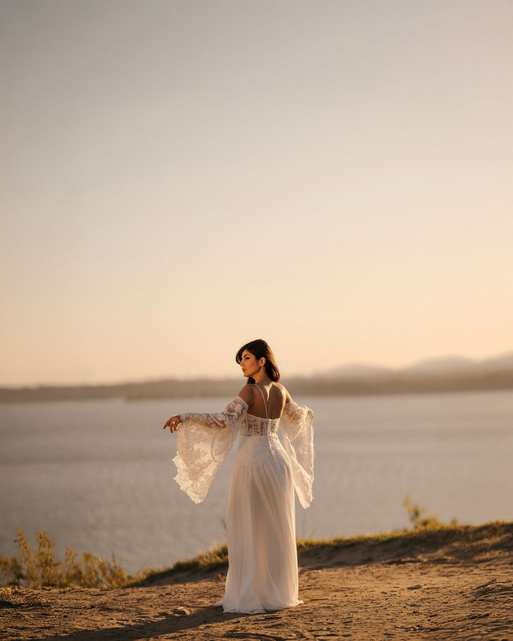 A person in a flowing white dress stands gracefully on a cliff overlooking a serene lake at sunset.
