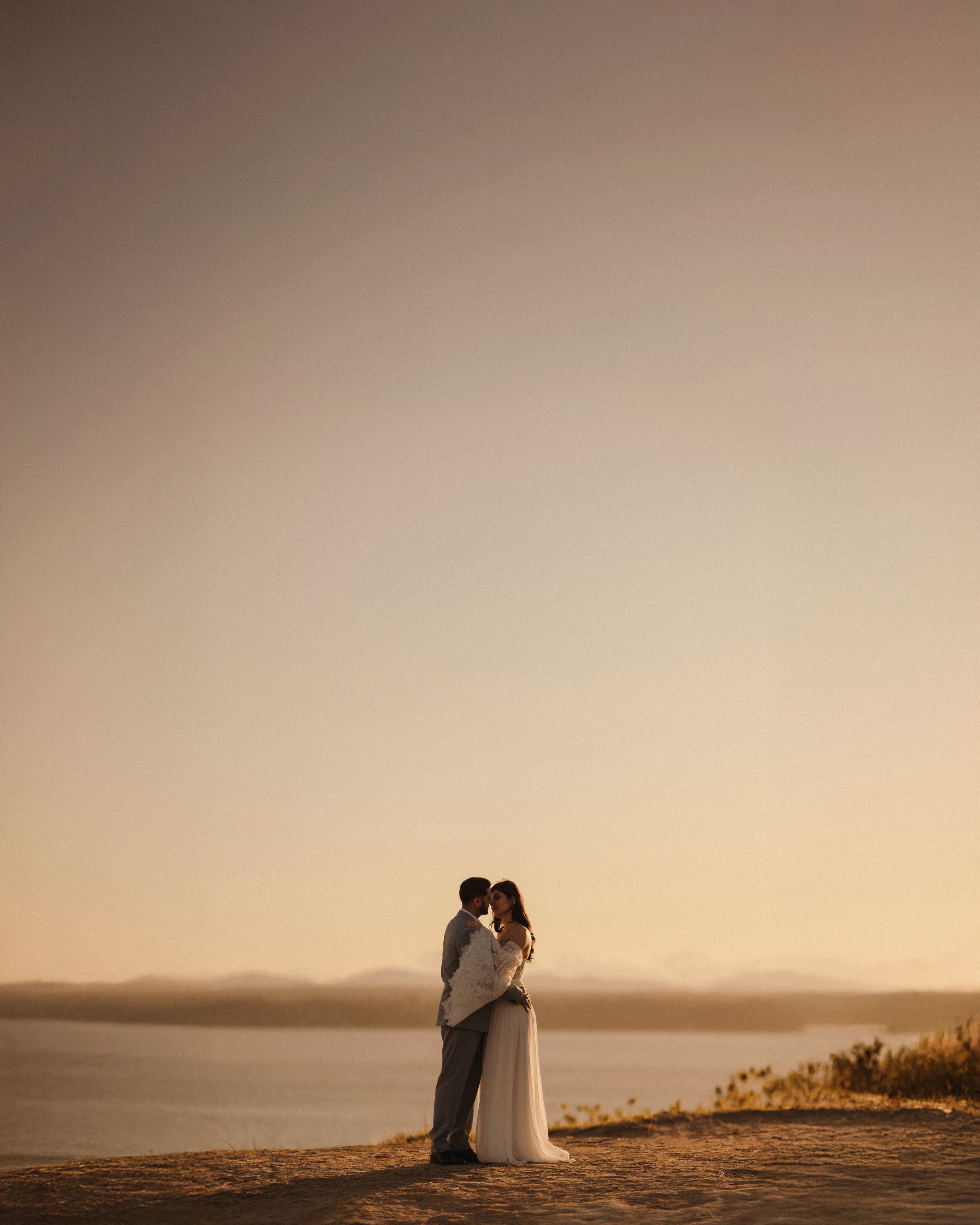 A couple in wedding attire silhouetted against a misty golden sunset on a hilltop overlooking a scenic landscape.
