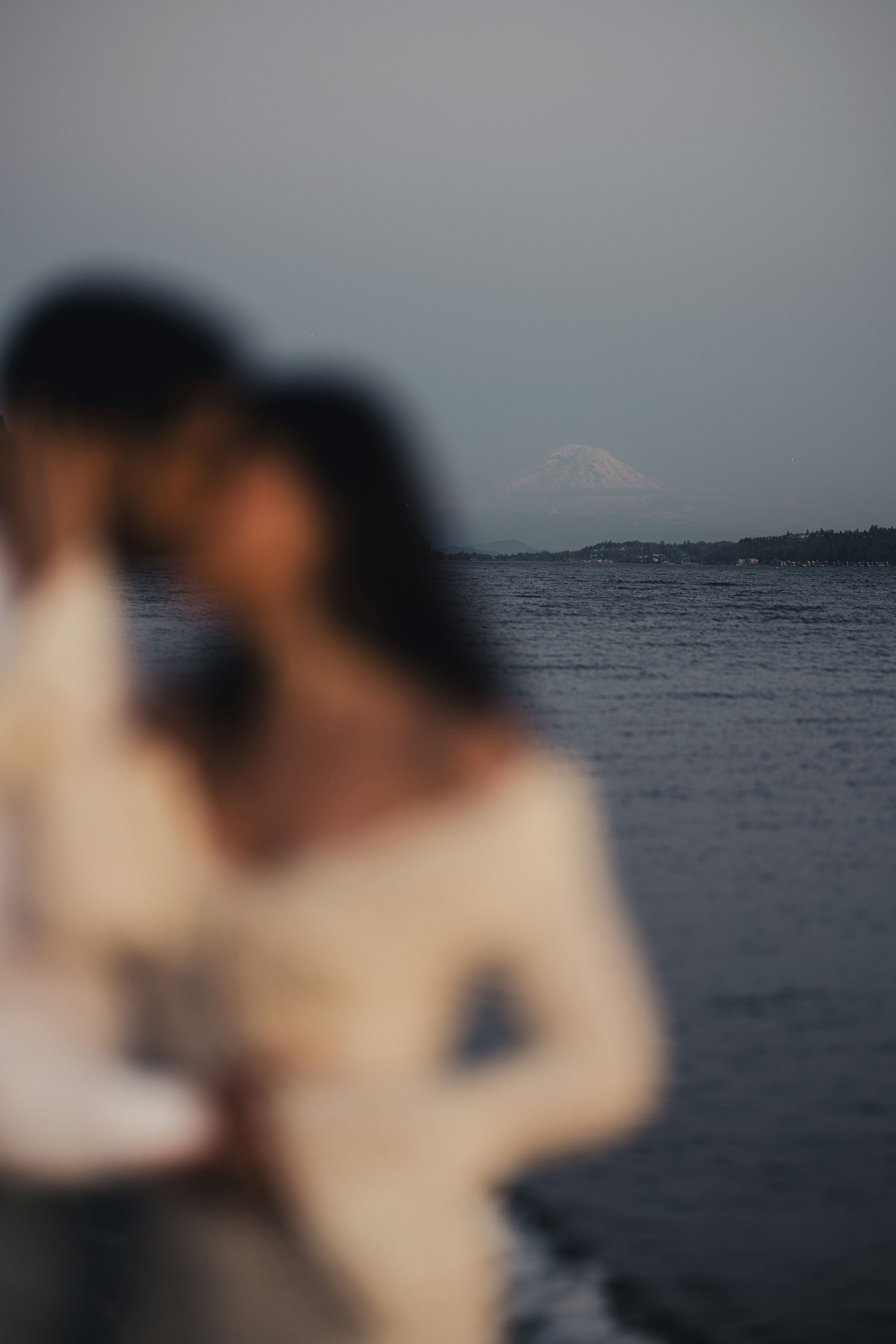 A romantic couple embraces against a moody ocean backdrop with mountains visible in the distance.
