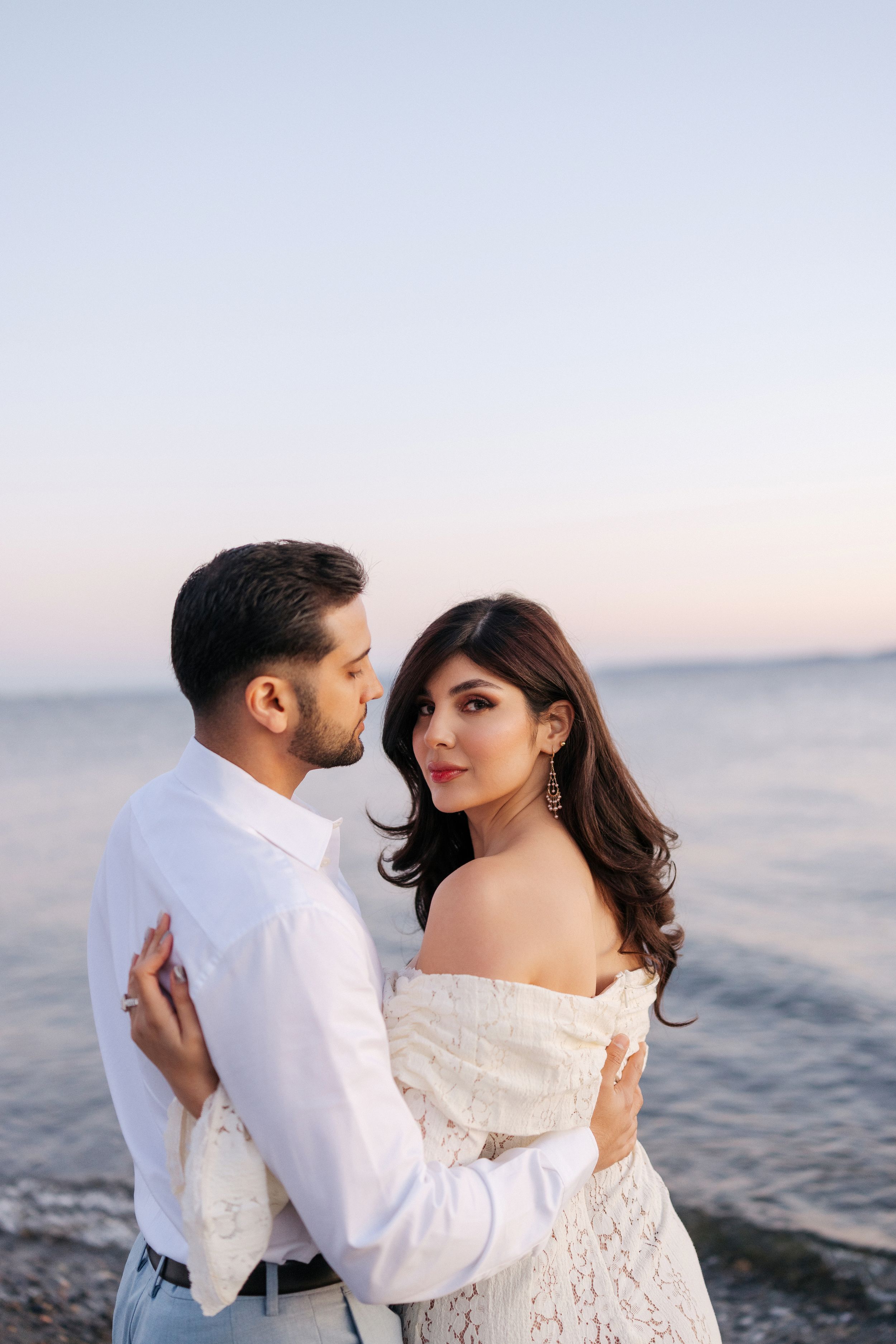 A romantic couple in white outfits embraces by the ocean during sunset in a series of intimate poses.