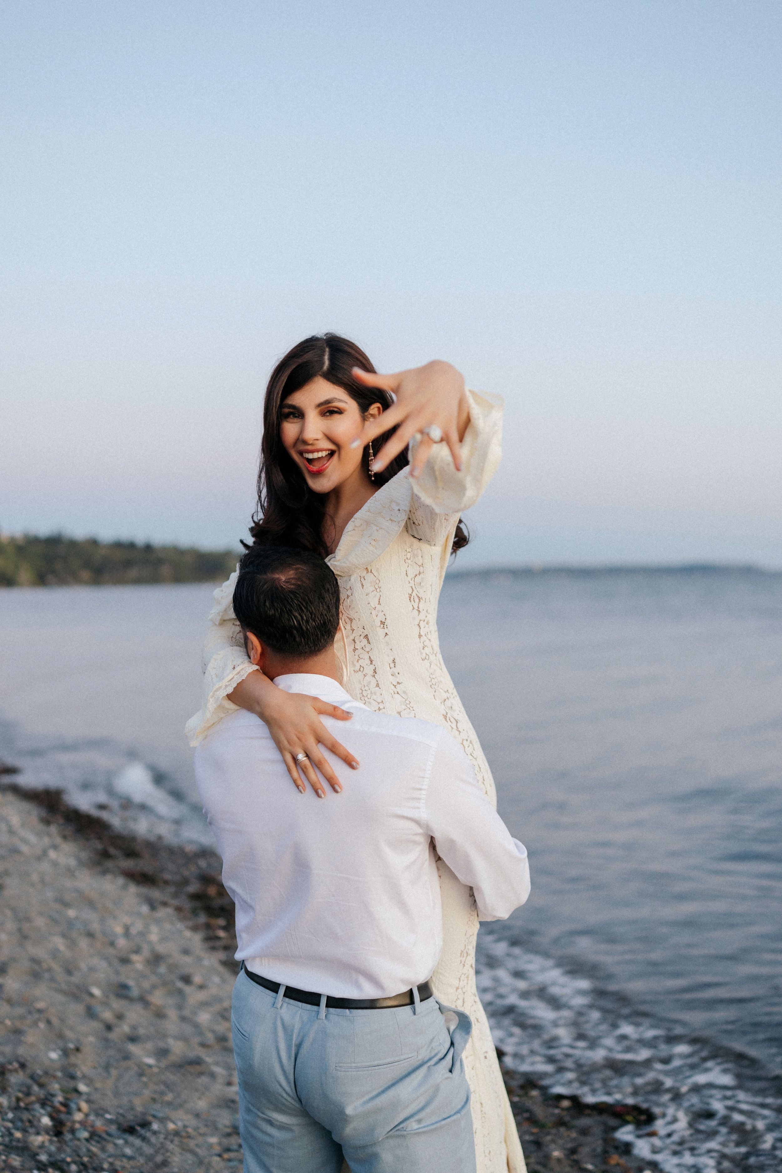 A couple embraces joyfully on a rocky beach with water and distant shoreline visible in the background.
