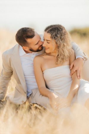 Romantic couple shares intimate moment in wheat field at sunset wearing light colored formal attire.