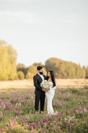 A couple shares a romantic moment in a field of pink wildflowers with golden trees in the background at sunset.