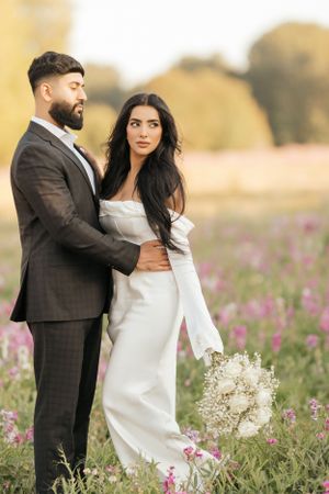 A couple poses together in a wildflower meadow at sunset, with the bride holding a delicate white bouquet.