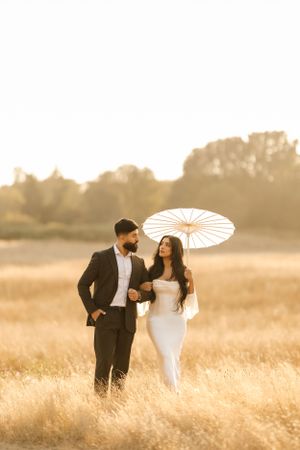 Couple in formal attire holding white parasol in golden wheat field during sunset with dreamy backlit atmosphere.
