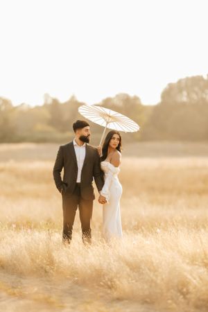 A couple in formal wedding attire stands in a golden field at sunset with a white parasol casting soft shadows.