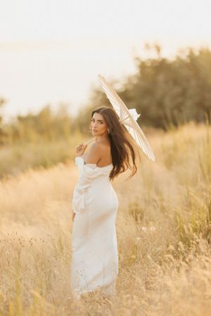 Ethereal outdoor portrait in a golden field at sunset with a white dress and delicate parasol creating a dreamy atmosphere.