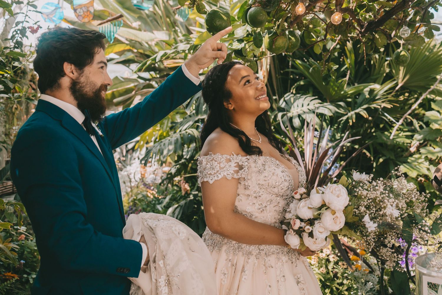 A couple wearing wedding attire are in a greenhouse and the groom is pointing up at a plant. Seymour Conservatory Tacoma