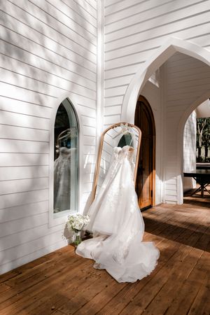 A flowing white wedding dress hangs elegantly in a rustic white chapel hallway with wooden floors and arched doorways.