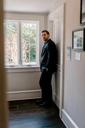 A groom in a dark suit leans against a doorframe in a bright hallway, hands in pockets, gazing at the camera.