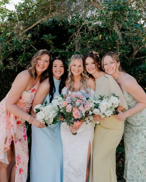 Bride in white lace gown poses with four bridesmaids in pastel dresses holding floral bouquets outdoors.