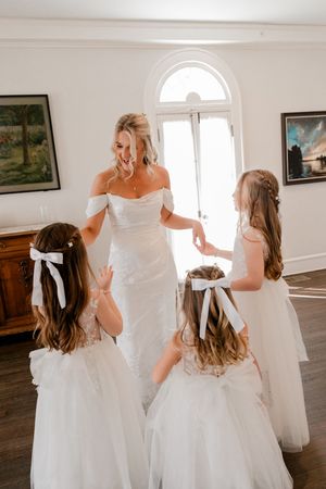 Bride in white off-shoulder gown laughing with three flower girls in white dresses with bow hair accessories indoors.