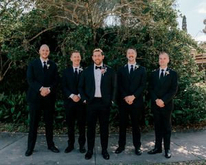 Groom in bow tie stands with four groomsmen in black tuxedos outdoors at a wedding ceremony.