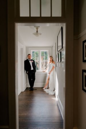 A bride and groom stand elegantly in a sunlit hallway, she in a white gown and he in a black tuxedo with bow tie.