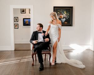 Elegant bride in white gown stands beside groom in tuxedo seated in chair inside a gallery with artwork on walls.