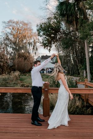 A groom twirls his bride in a flowing white dress on a wooden deck overlooking a pond at their outdoor wedding reception.