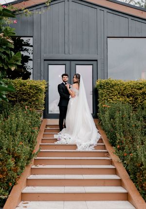 A couple in wedding attire stands in front of a gray building with wooden steps and green hedges on a cloudy day.