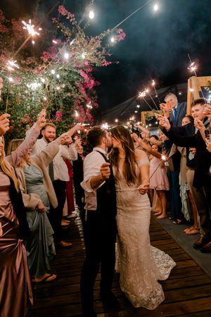 Wedding guests celebrate with sparklers on a wooden deck during an outdoor evening reception under string lights.