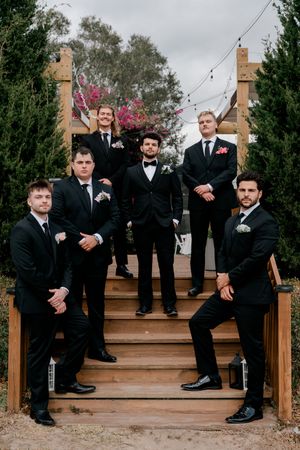 Groomsmen in black suits pose casually on wooden stairs outdoors during wedding photoshoot.