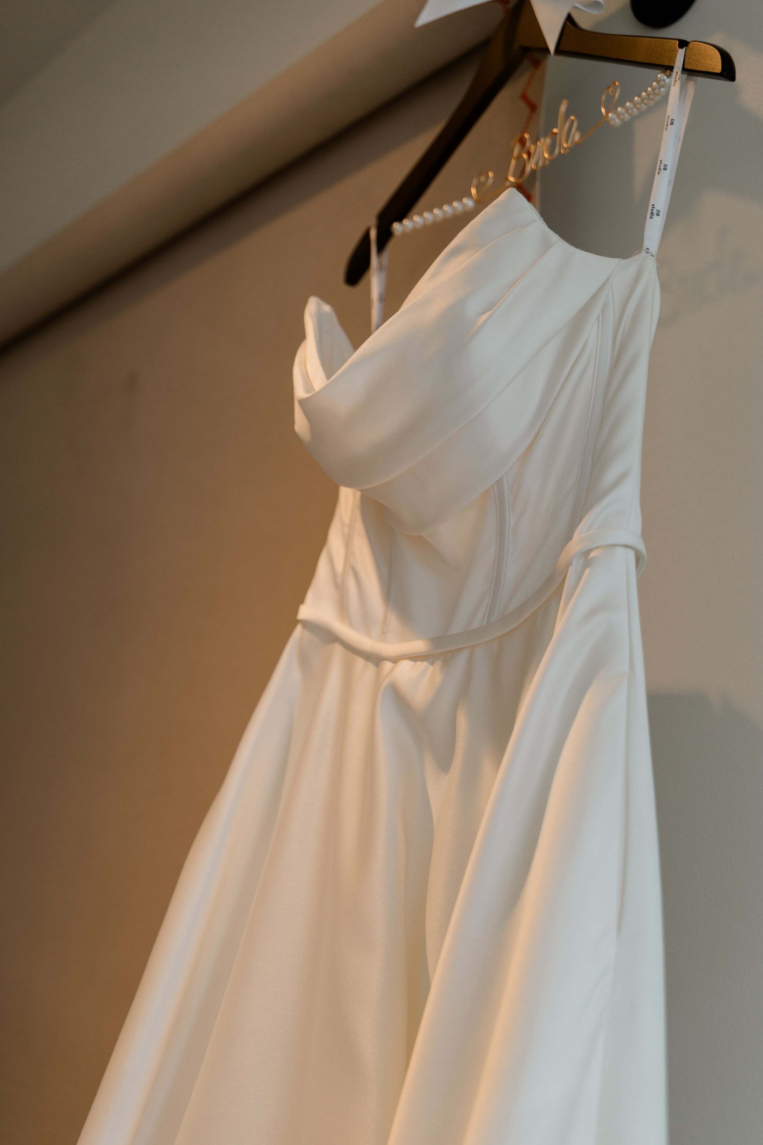 White wedding gown hanging on a decorative bride hanger with pearl details against a neutral wall.