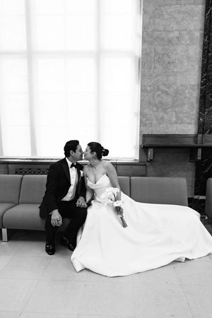 Black and white photo of a bride and groom sharing a kiss while seated on a couch near a large window.