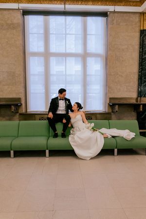 Bride and groom sit together on a green bench in an elegant hall, gazing at each other lovingly on their wedding day.