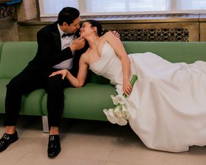 Bride and groom sharing a romantic kiss on a green velvet sofa, bride in strapless white gown holding calla lilies.