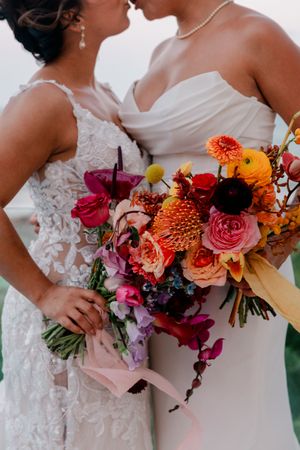 Two brides in white gowns share a romantic moment holding a vibrant bouquet of colorful flowers.