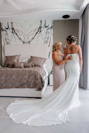 A bride in a white gown with train gets help from her bridesmaid in a neutral bedroom setting.